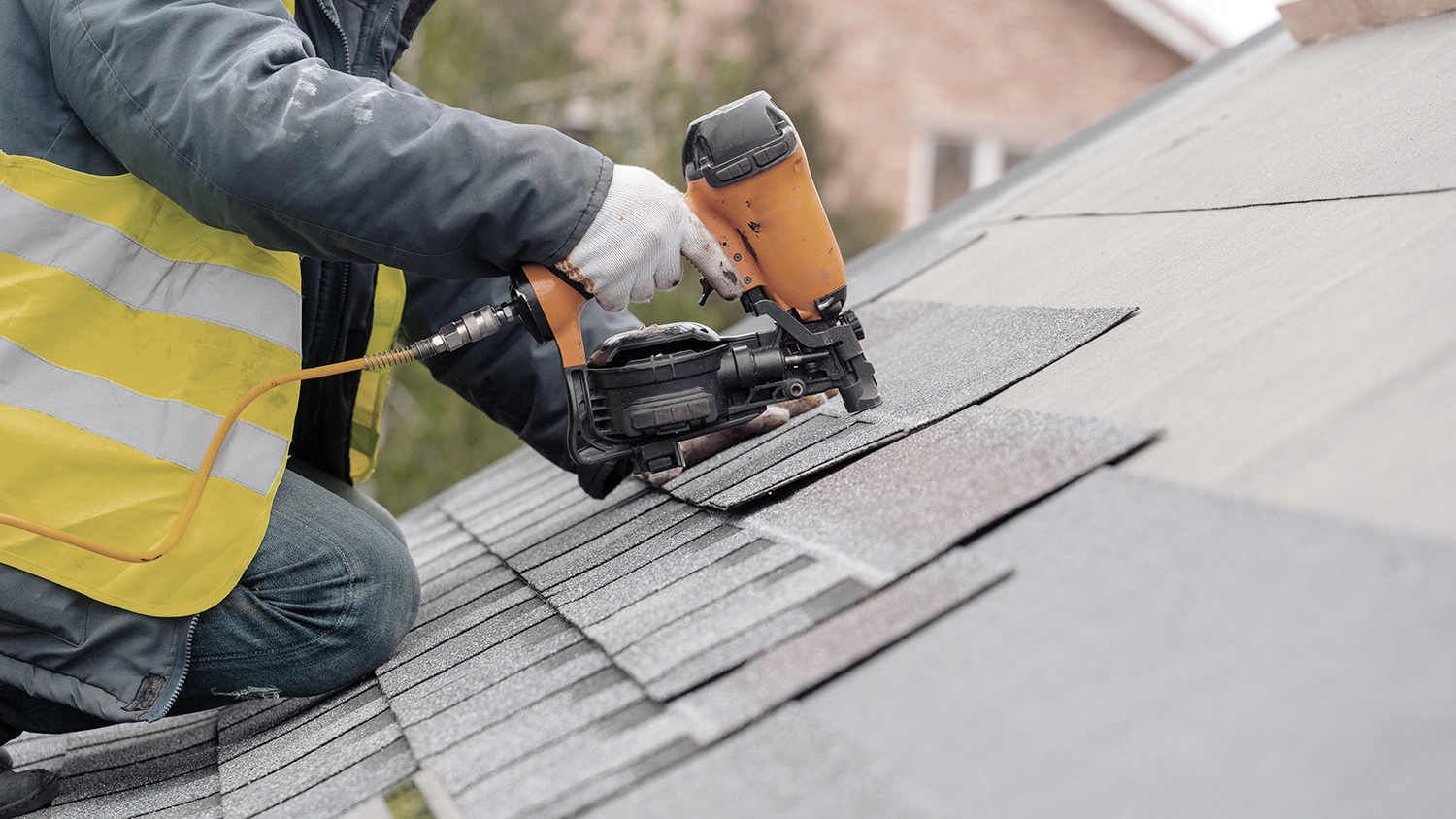 Roofer worker in special protective work wear and gloves, using air or pneumatic nail gun and installing asphalt or bitumen shingle on top of the new roof under construction residential building